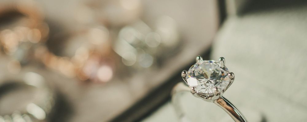 En primer plano, un anillo de compromiso con un diamante de corte brillante dentro de una caja, con otros anillos desenfocados al fondo.