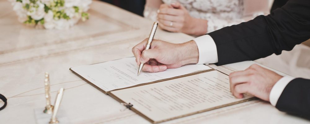 Novio y novia firmando la documentación oficial de su matrimonio, con un elegante ramo de flores de fondo.