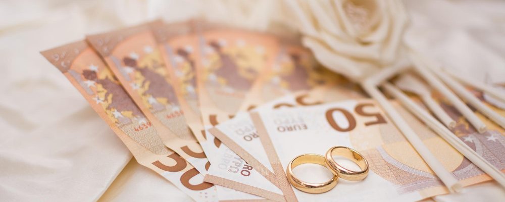 Dos anillos de boda de oro sobre billetes de 50 euros, junto a flores decorativas en un fondo claro.