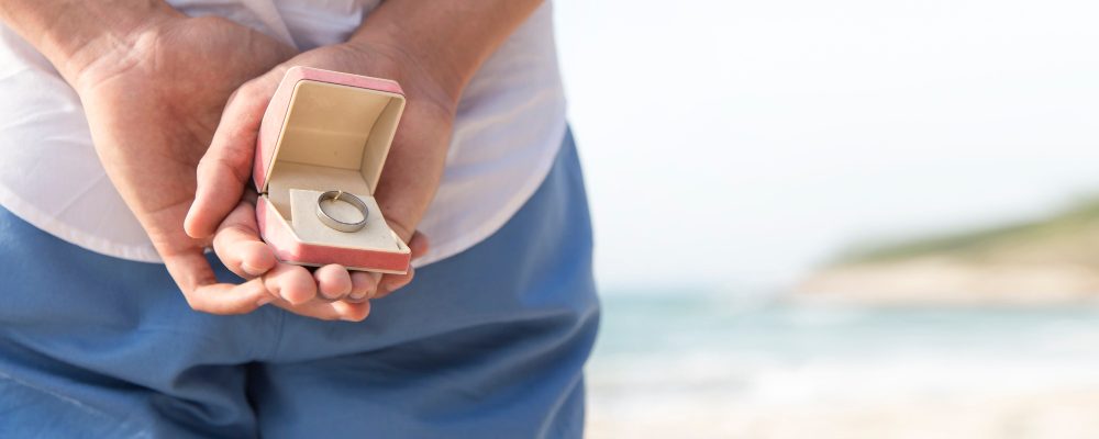 Persona sosteniendo una caja con un anillo de compromiso en la playa, listo para hacer una propuesta de matrimonio.